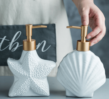 White ceramic soap dispensers in starfish and shell shape with gold-tone pumps, a hand pressing the shell pump