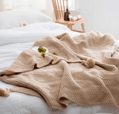 Beige chunky knit throw spread on a bed with pompoms and small fruit on a plate