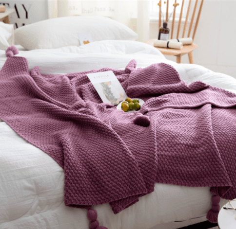 Plum chunky knit throw laid on a bed with a card and olives, pompoms at corners
