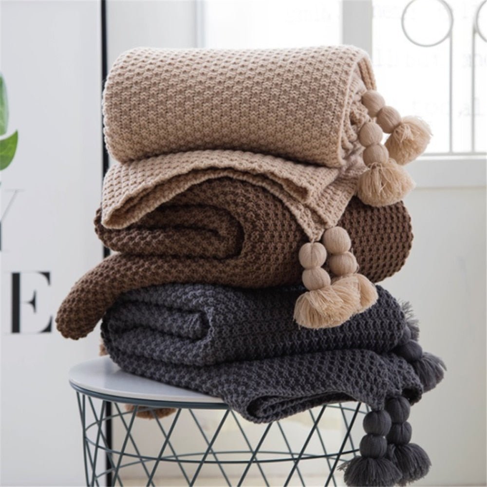 Chunky knit throw in beige, brown, dark grey and plum stacked on a stool with pompom tassels visible