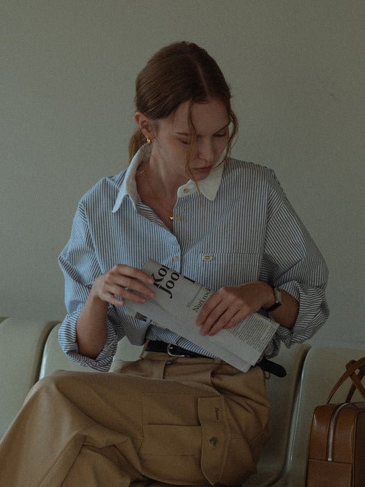 Model seated on a cream sofa in the striped shirt with rolled sleeves, reading a newspaper