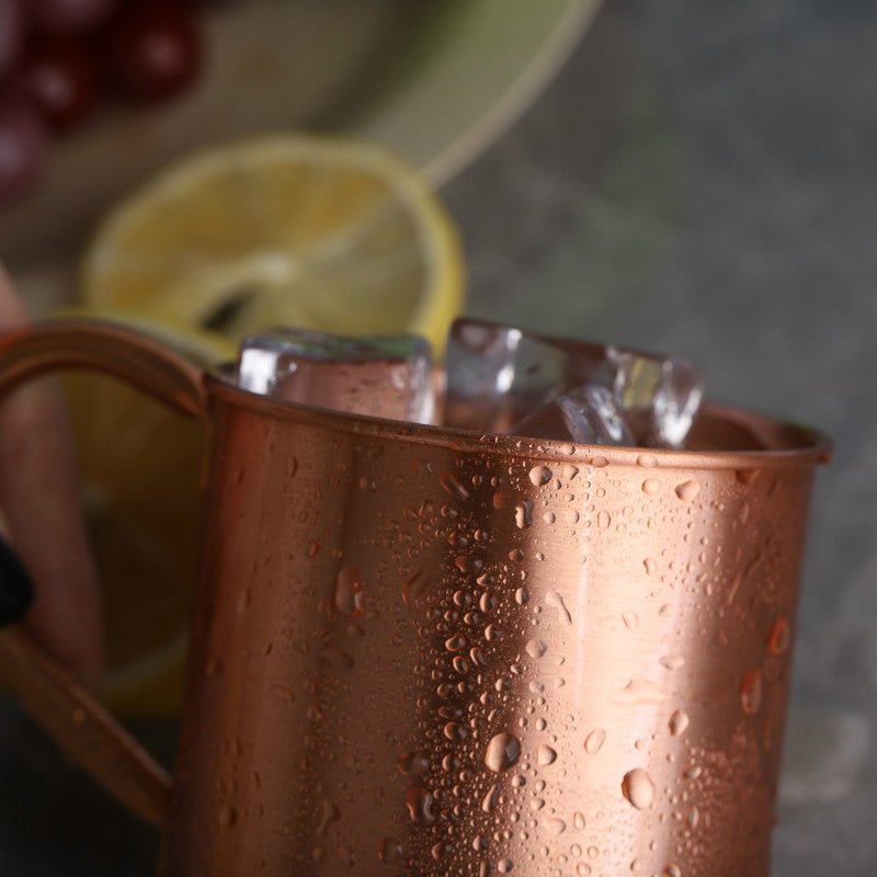 Copper cocktail mug with handle, filled with ice, condensation visible, lemon slice and grapes in background