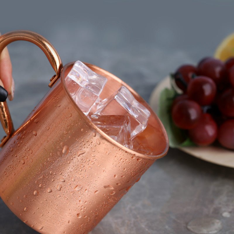 Copper cocktail mug with handle, filled with ice, held in hand, condensation visible, grapes and lemon in background