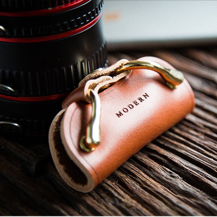Brown cowhide key organiser with two gold-tone clips on a wooden table next to a camera lens