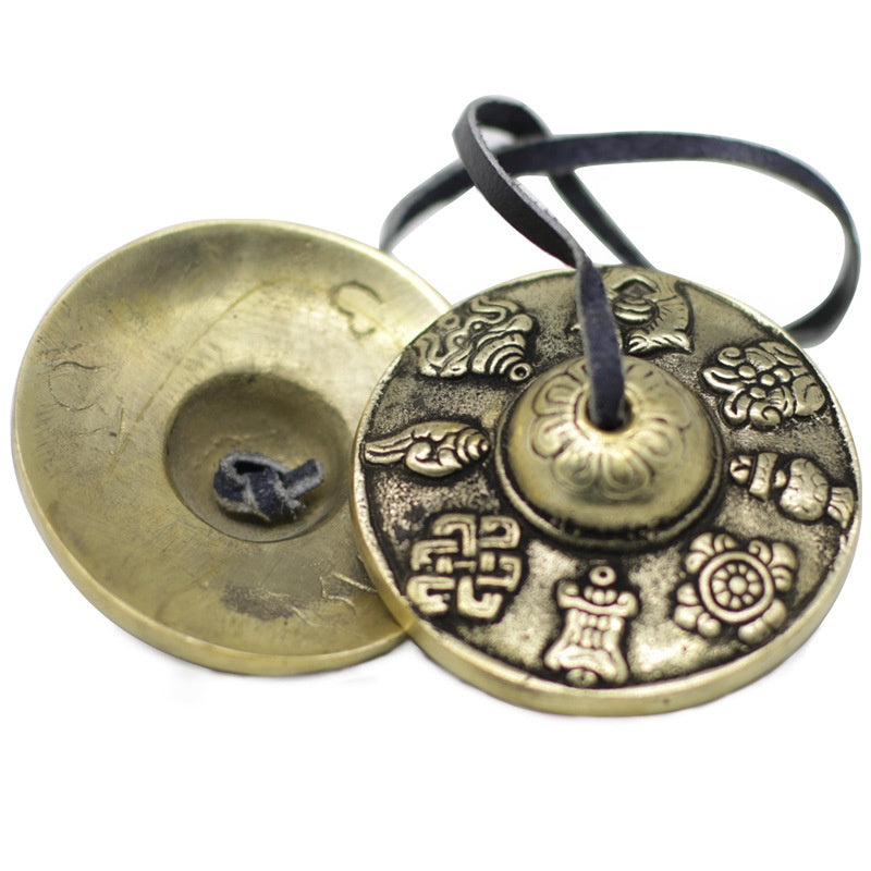 Tingsha cymbal pair with eight symbols, brass coloured, detailed embossed pattern, joined by black cord, flat lay on white