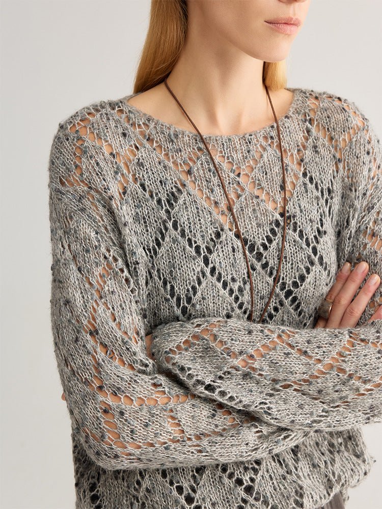 Close-up of a grey fleck open-knit jumper with a diamond pattern, arms crossed, long pendant necklace