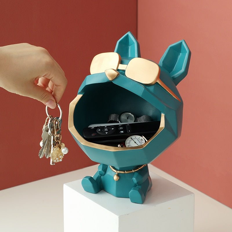 Teal dog figure catch-all with gold-tone sunglasses, a remote control and a watch in its open mouth, a hand holding keys beside it