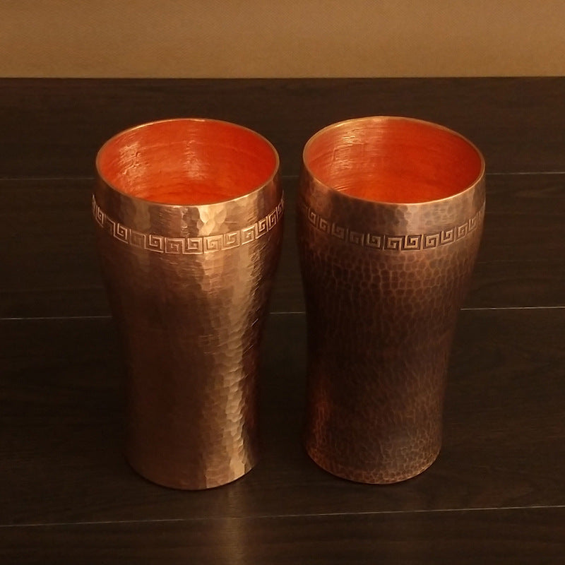 Two hammered copper beer tumblers with Greek-key border, curved style, on dark surface