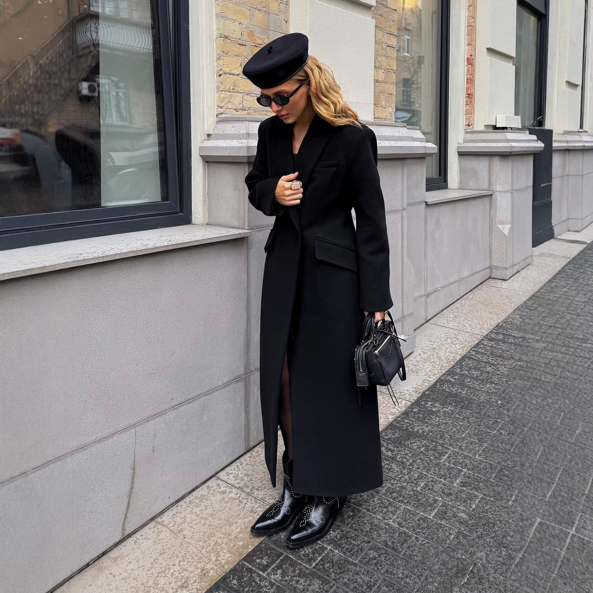 Floor-length single-breasted tailored coat in black, front view of model standing on sidewalk, holding a bag