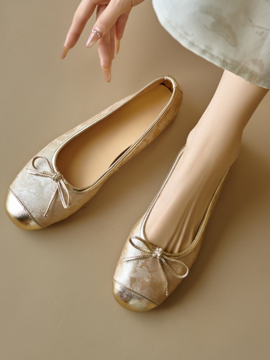 Gold floral ballet flats with metallic toe cap and bow, one shoe worn, one beside foot