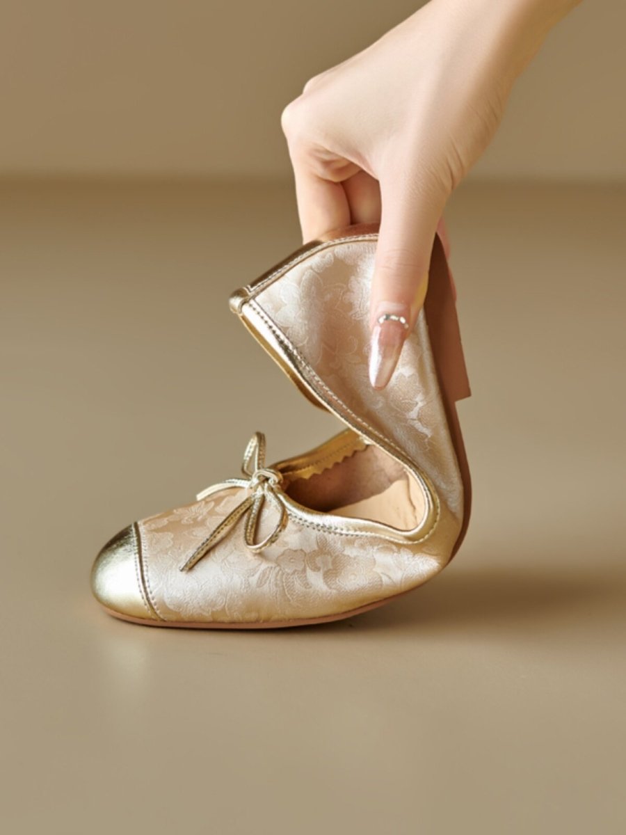 Gold floral ballet flats with metallic toe cap and bow, held by hand, close-up detail