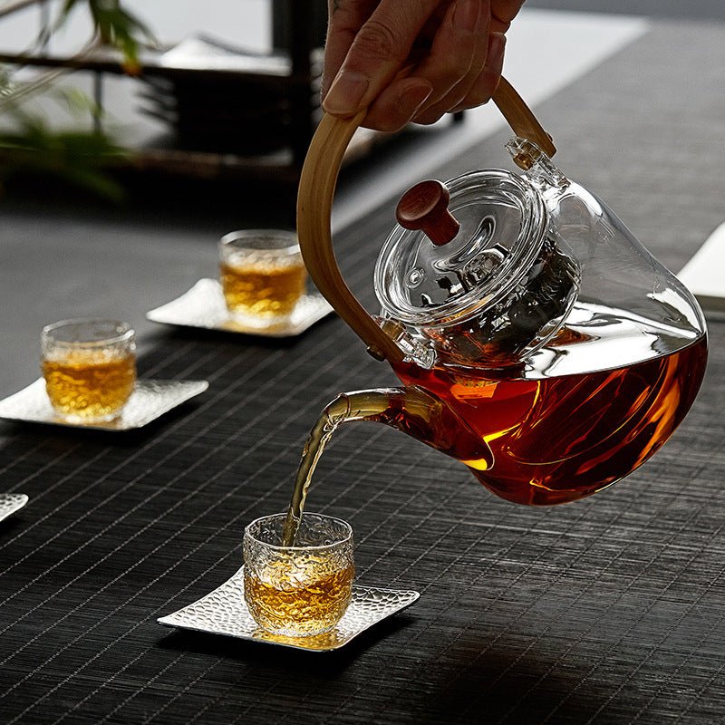 Glass teapot pouring tea into a small cup, in use on a dark table with cups and coasters, side view