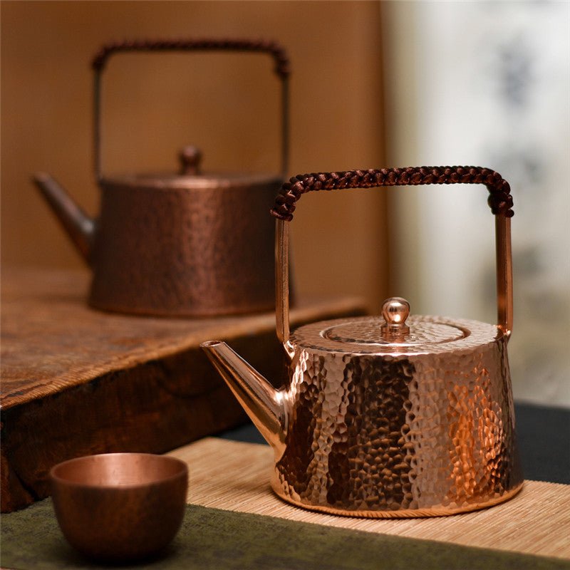 Hammered copper teapot with braided handle, front view on wooden surface, small cup nearby
