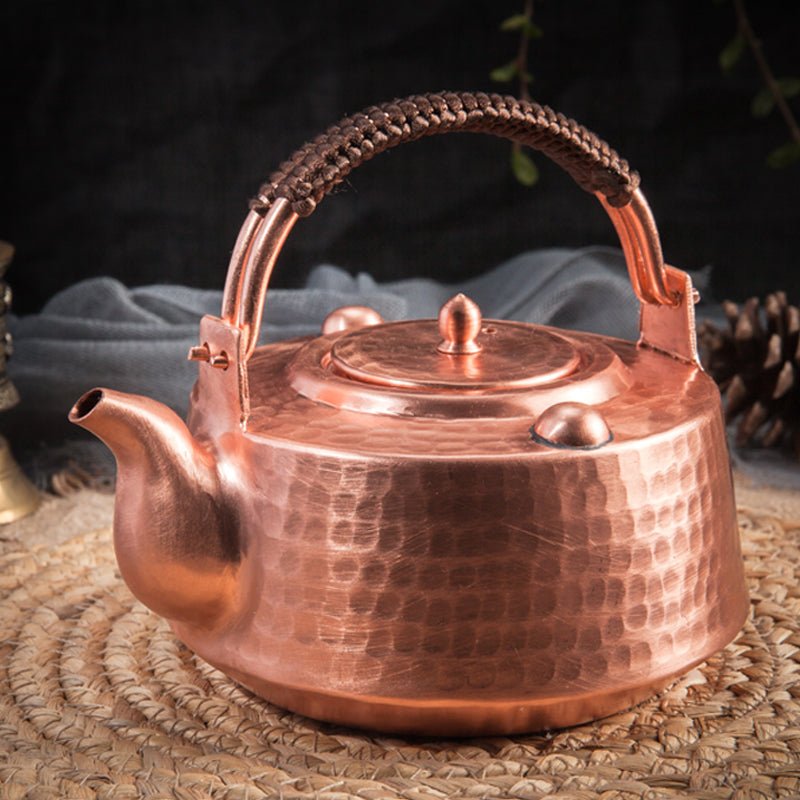 Hammered copper teapot with cord-wrapped top handle, shown on a woven mat with dark background