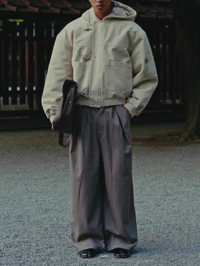 The hooded jacket worn outdoors on gravel, hand in the pocket of wide brown trousers