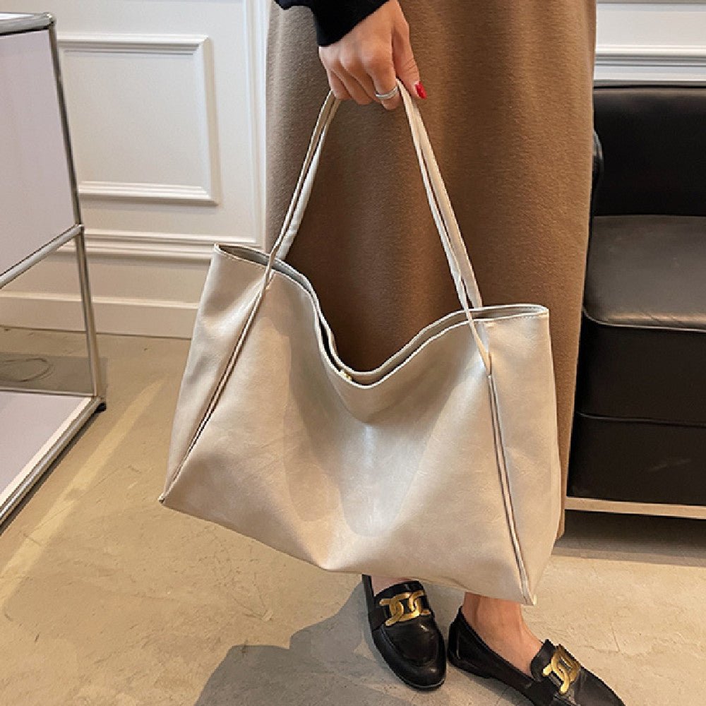 Large off-white faux leather tote bag held by its two long straps, next to a brown skirt and black loafers