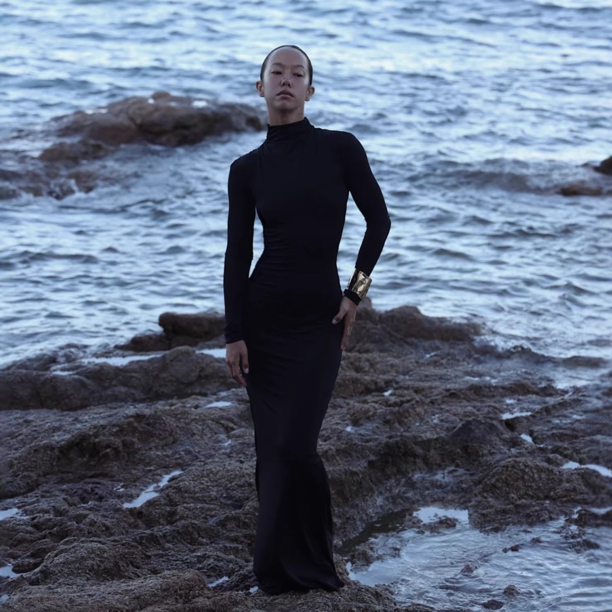 Woman in a black floor-length turtleneck maxi dress with long sleeves standing on rocks by the sea