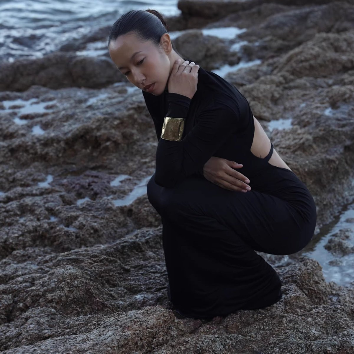 Model crouching on rocks in the black open-back dress, wearing a gold cuff