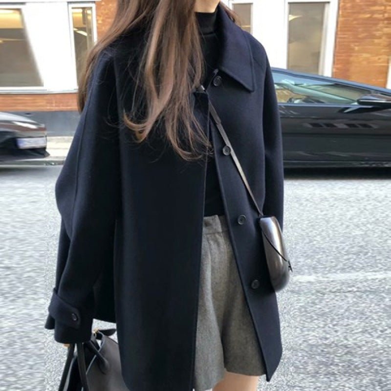Woman in the navy oversized coat, worn open over grey shorts with a small shoulder bag