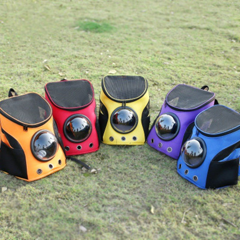 Five pet carrier backpacks with bubble windows standing on grass in orange, red, yellow, purple and blue