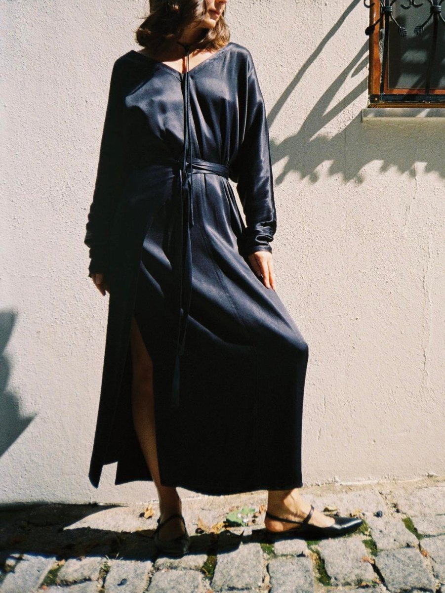 Navy maxi dress in the sun against a white wall, slit open at the front, black flat shoes