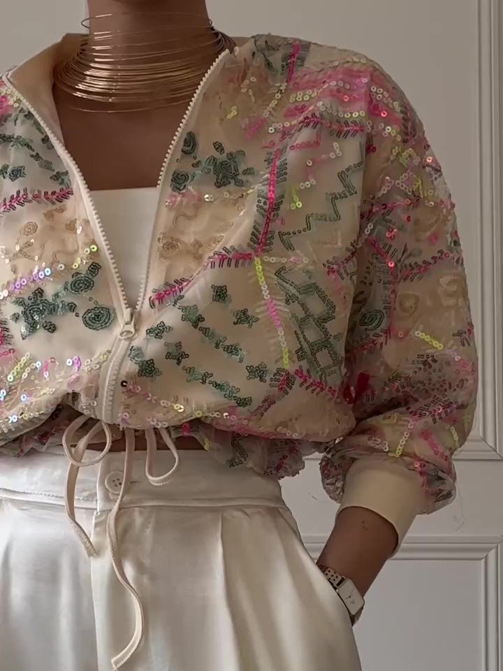 Sheer cream cropped jacket with pink, green and gold sequin embroidery, zipped, over cream trousers