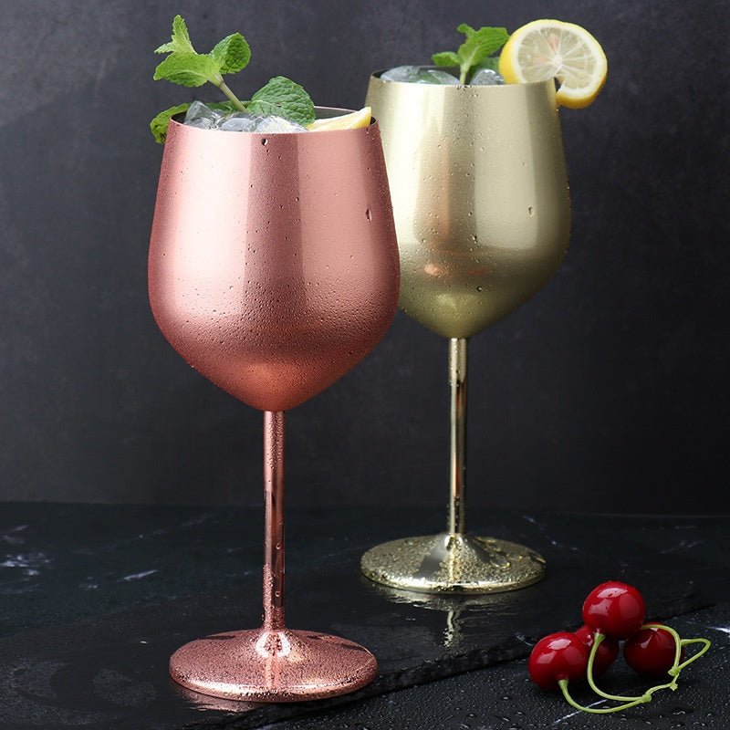 Stainless steel wine glass in rose gold and gold, filled with ice, lemon slice, and mint, on dark surface with cherries