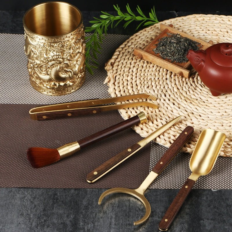 Tea ceremony tools with brass-coloured metal and red-brown wooden handles, laid flat on a textured mat near a teapot and tea