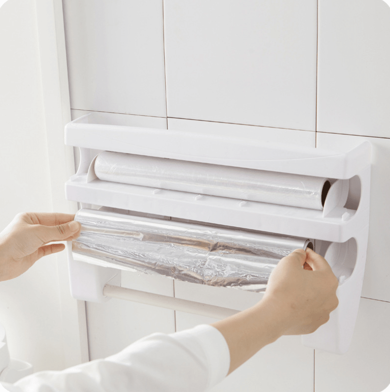 White wall-mounted holder on tiles, two hands pulling a sheet of foil out of the lower compartment